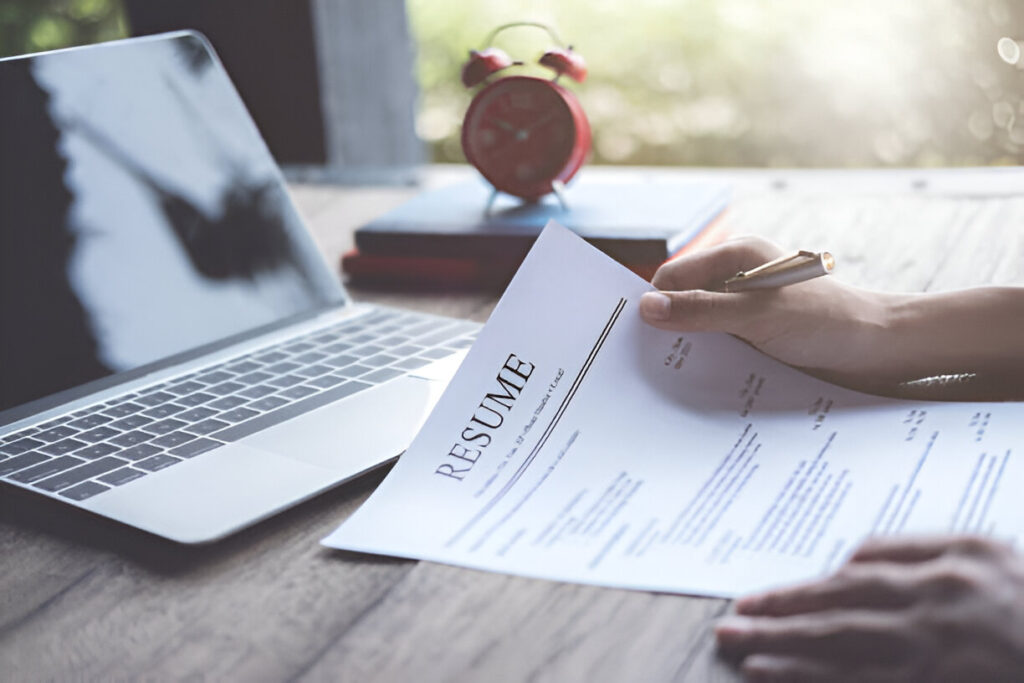 Hand holding a pen over a resume beside a laptop.